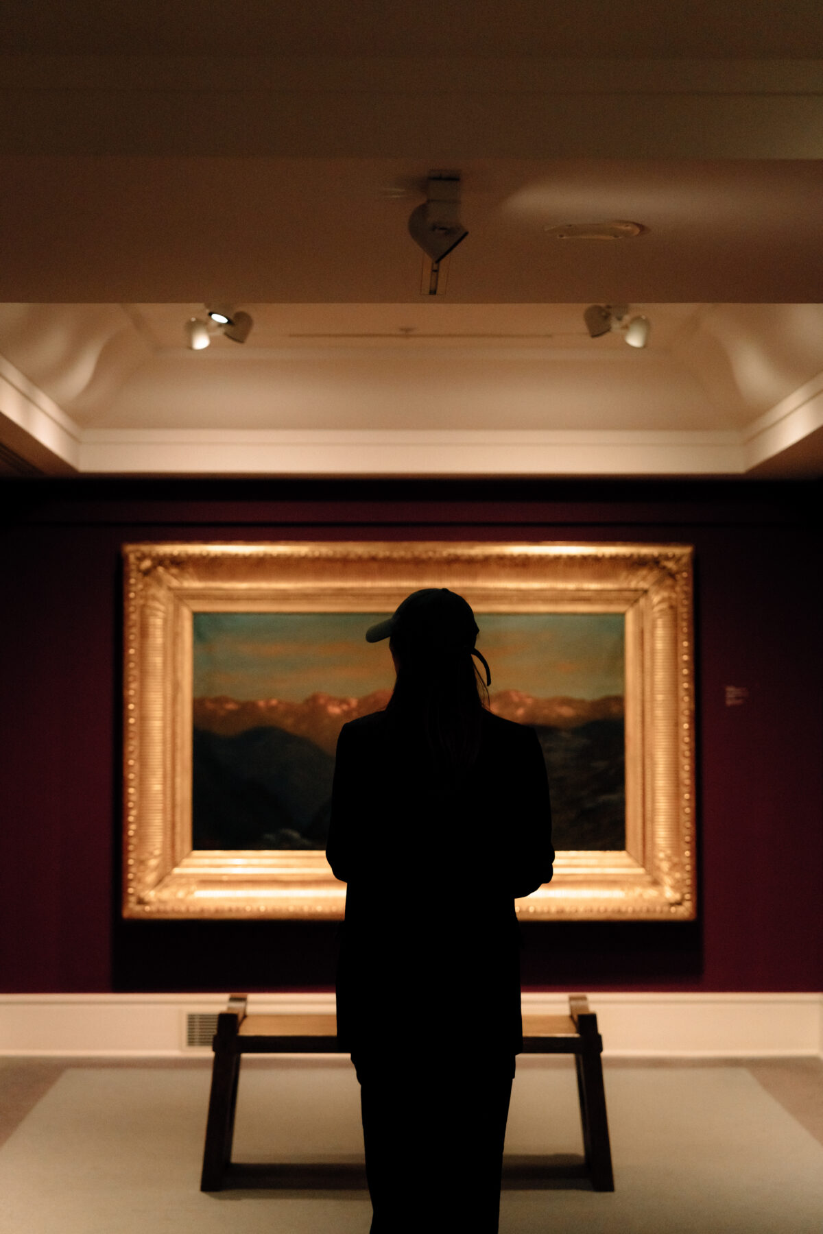 A silhouetted person wearing a cap stands facing a large, framed landscape painting in a softly lit art gallery.