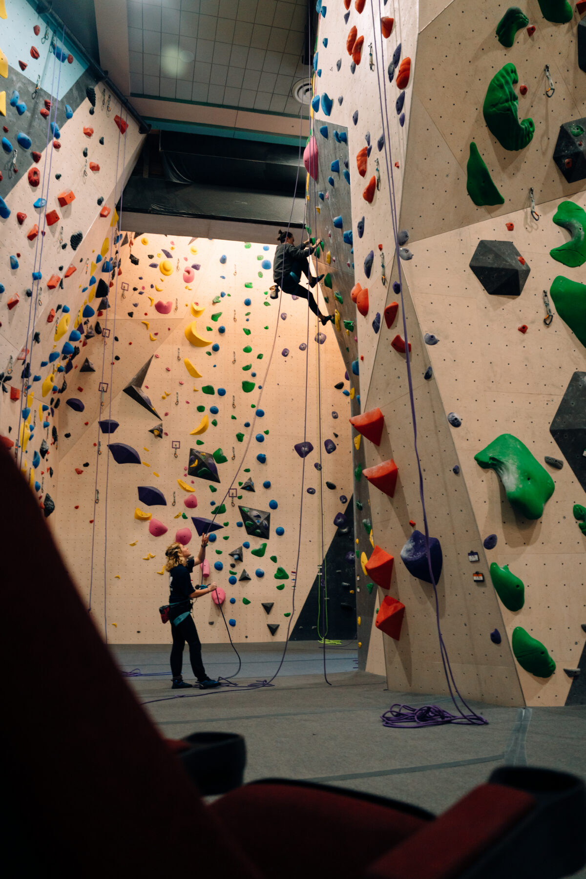 Climber on a tall indoor wall at East Peak Climbing with colourful routes and a belayer below.