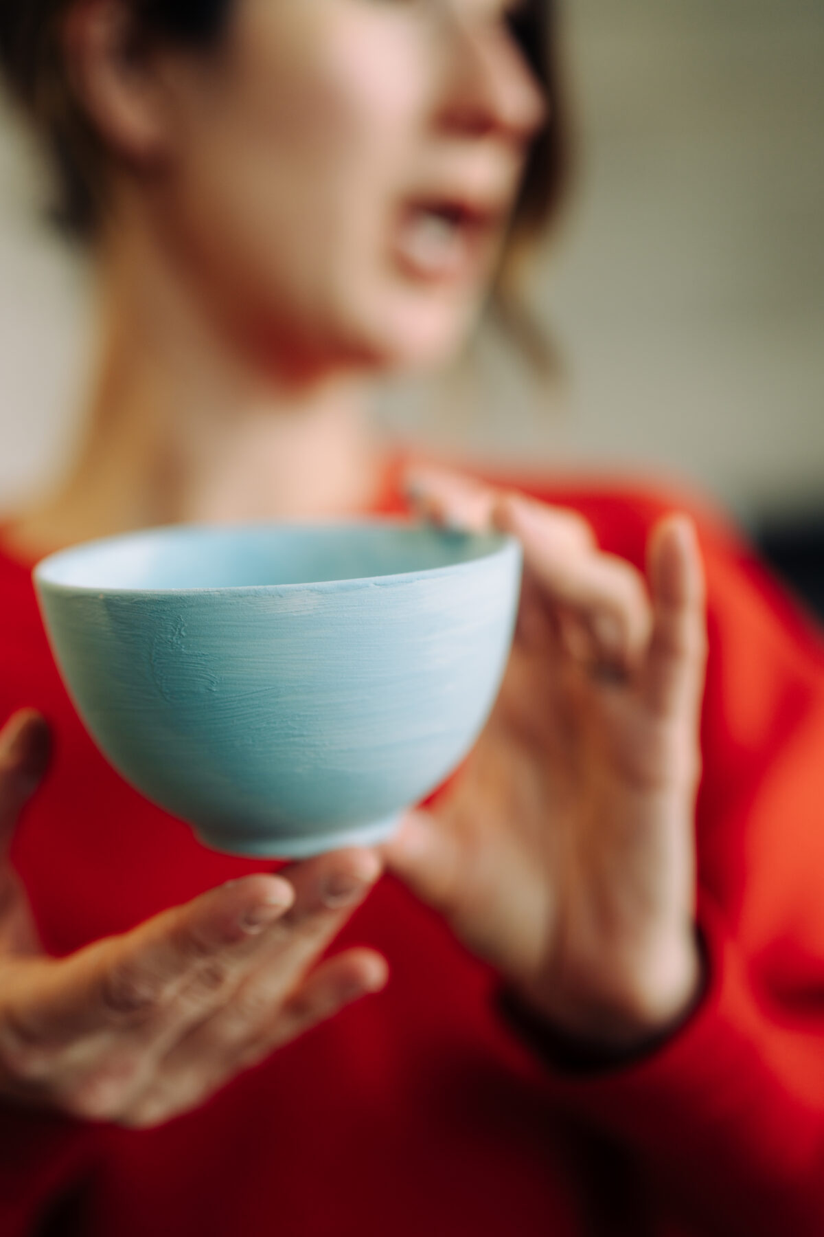 A close-up, softly blurred image of a person in a red top holding a small, light blue ceramic bowl with both hands, appearing to speak or gesture while presenting it.