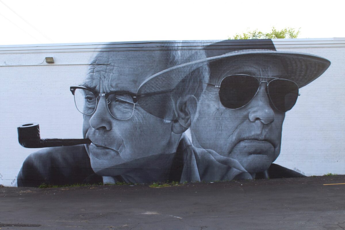 A large black-and-white mural depicting two men in profile, one wearing glasses and a pipe and the other in sunglasses and a hat, painted on an exterior wall.