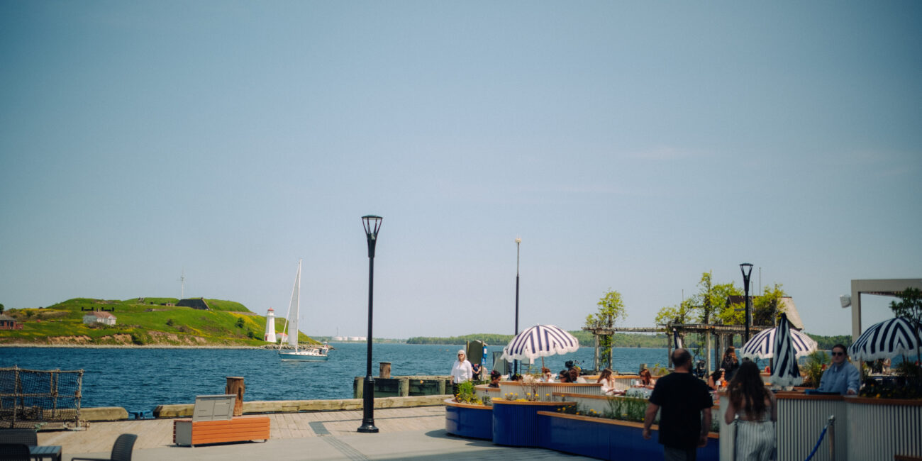 Halifax Waterfront boardwalk with harbour views, patio seating, and a sailboat passing Georges Island.