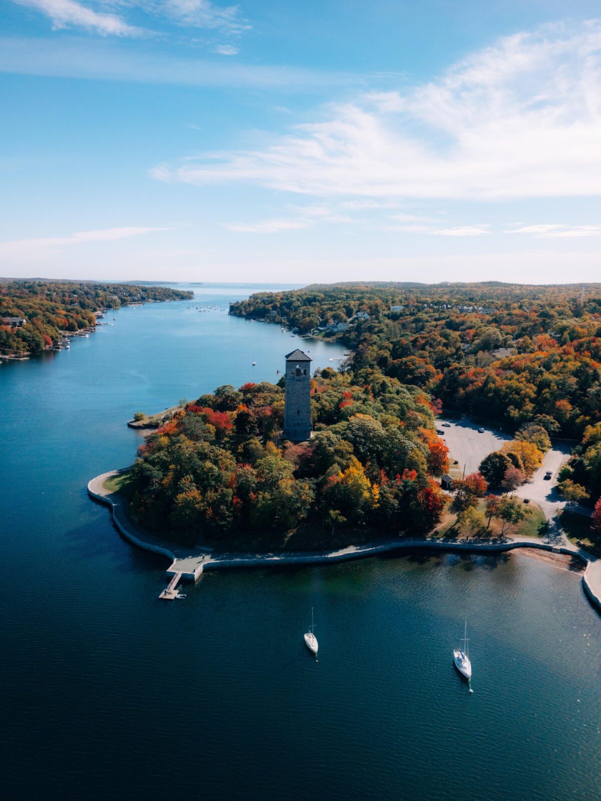 Aerial view of Sir Sandford Fleming Park in Halifax, showing a small tree-covered peninsula in autumn with vibrant red, orange, and green foliage, surrounded by calm blue water. The Dingle Tower stands prominently at the center, with a walking path tracing the shoreline, a small dock extending into the water, and a few sailboats anchored nearby under a clear sky.