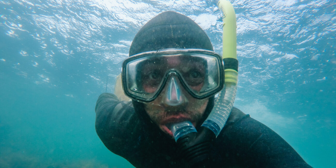 Underwater close-up of Saltwater Sean snorkeling in clear coastal water, wearing a black wetsuit, mask, and snorkel, looking directly at the camera with the water’s surface visible above.