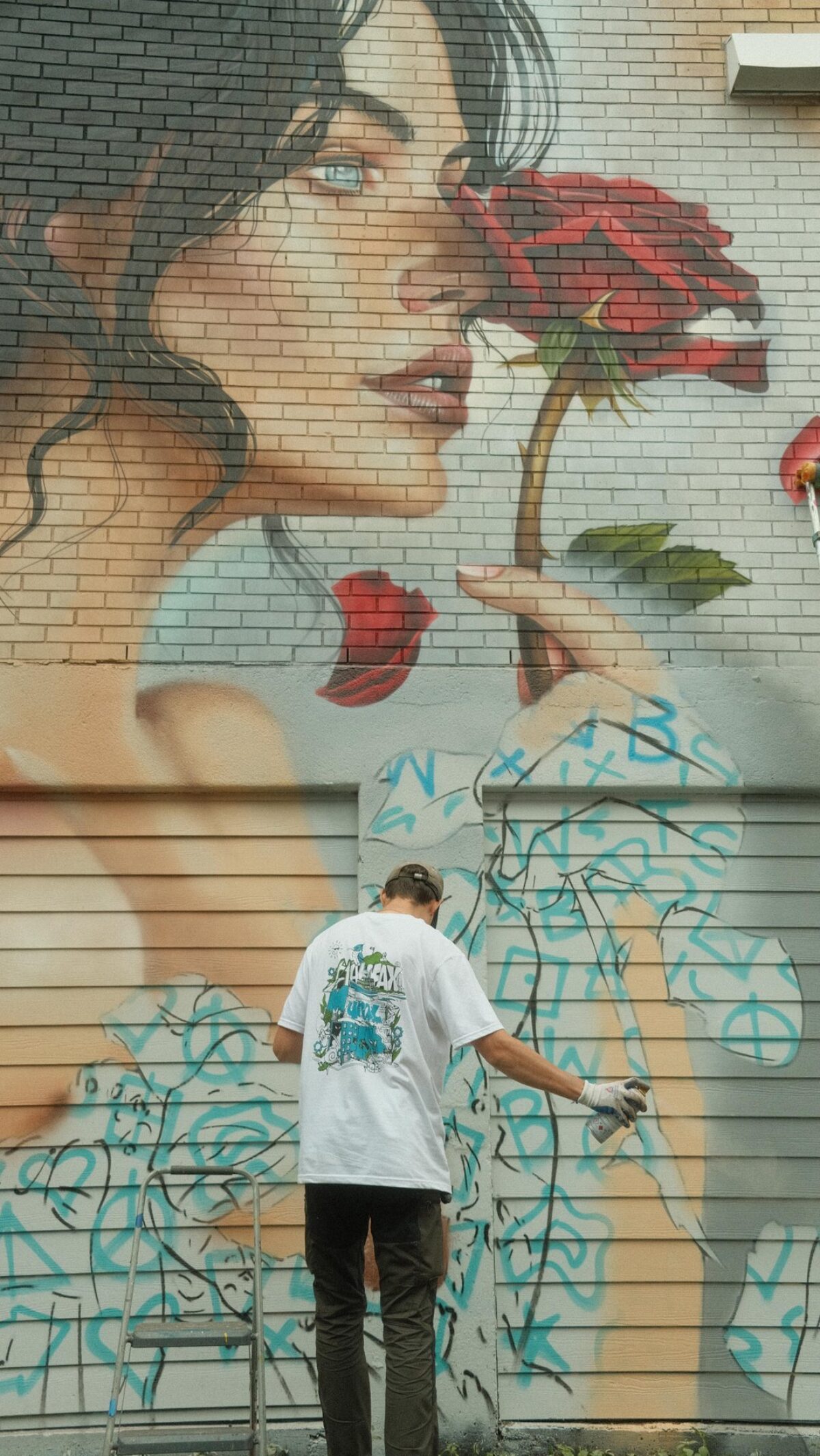 A large mural on a brick wall depicting a woman in profile holding a red rose near her face, with a person below spray-painting abstract blue shapes on the lower portion of the wall.