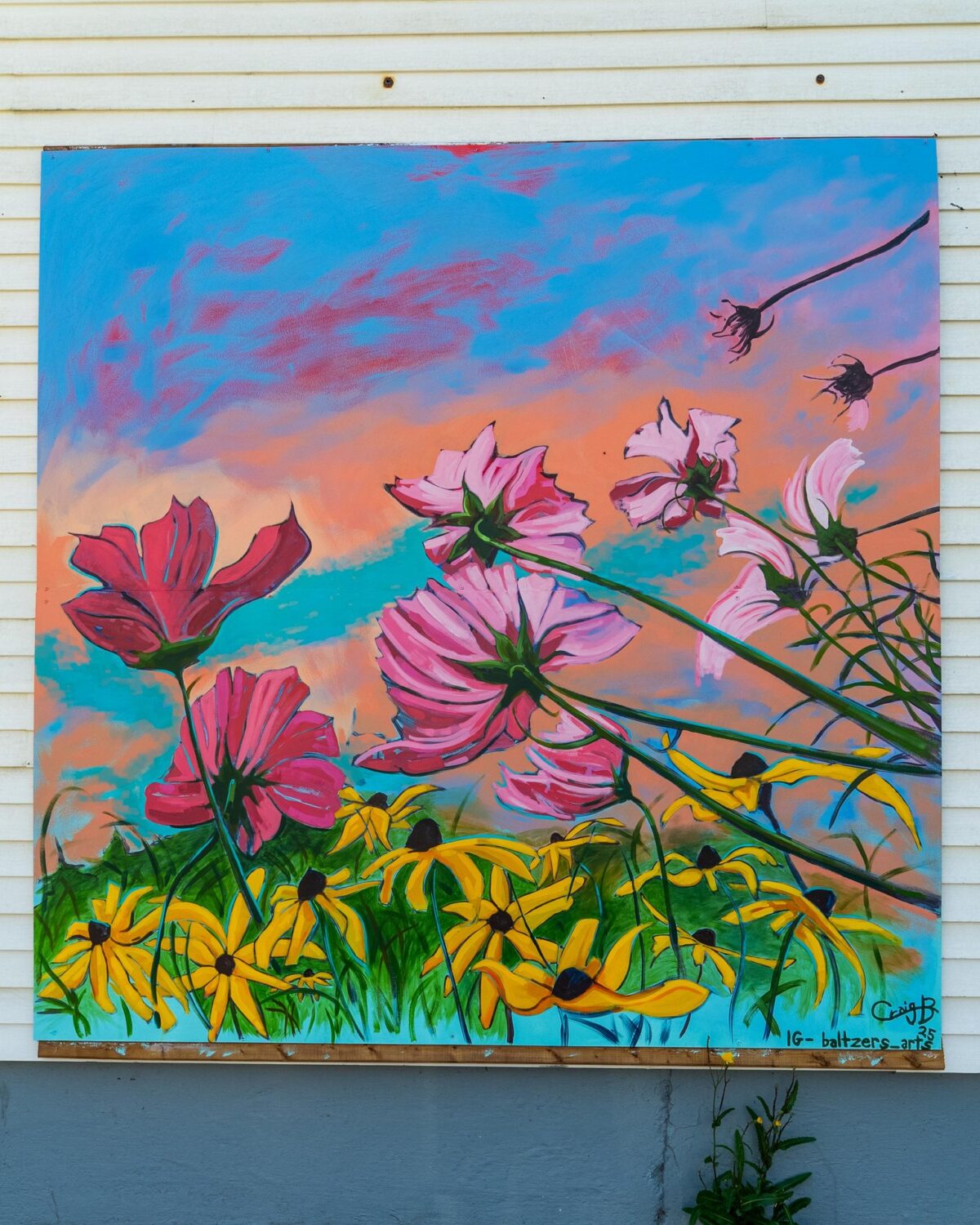 A vibrant mural of pink and red flowers with yellow blooms in the foreground, set against a colorful sky of blue, orange, and pink hues, painted on an exterior wall.