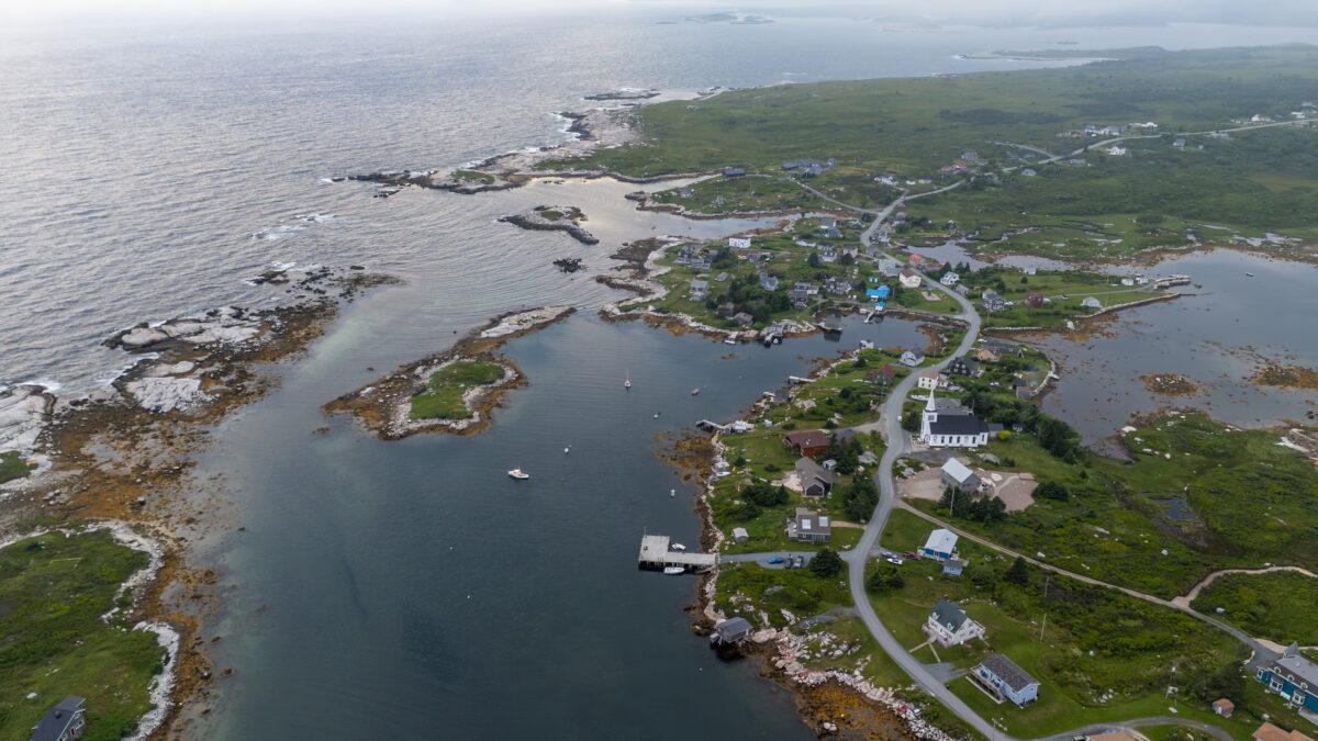 Aerial view of Prospect, Nova Scotia, showing a coastal fishing village with scattered houses, winding roads, and small islands surrounded by the Atlantic Ocean. Rocky shorelines and calm inlets frame the community, with boats in the water and low, green coastal landscape stretching inland under an overcast sky.