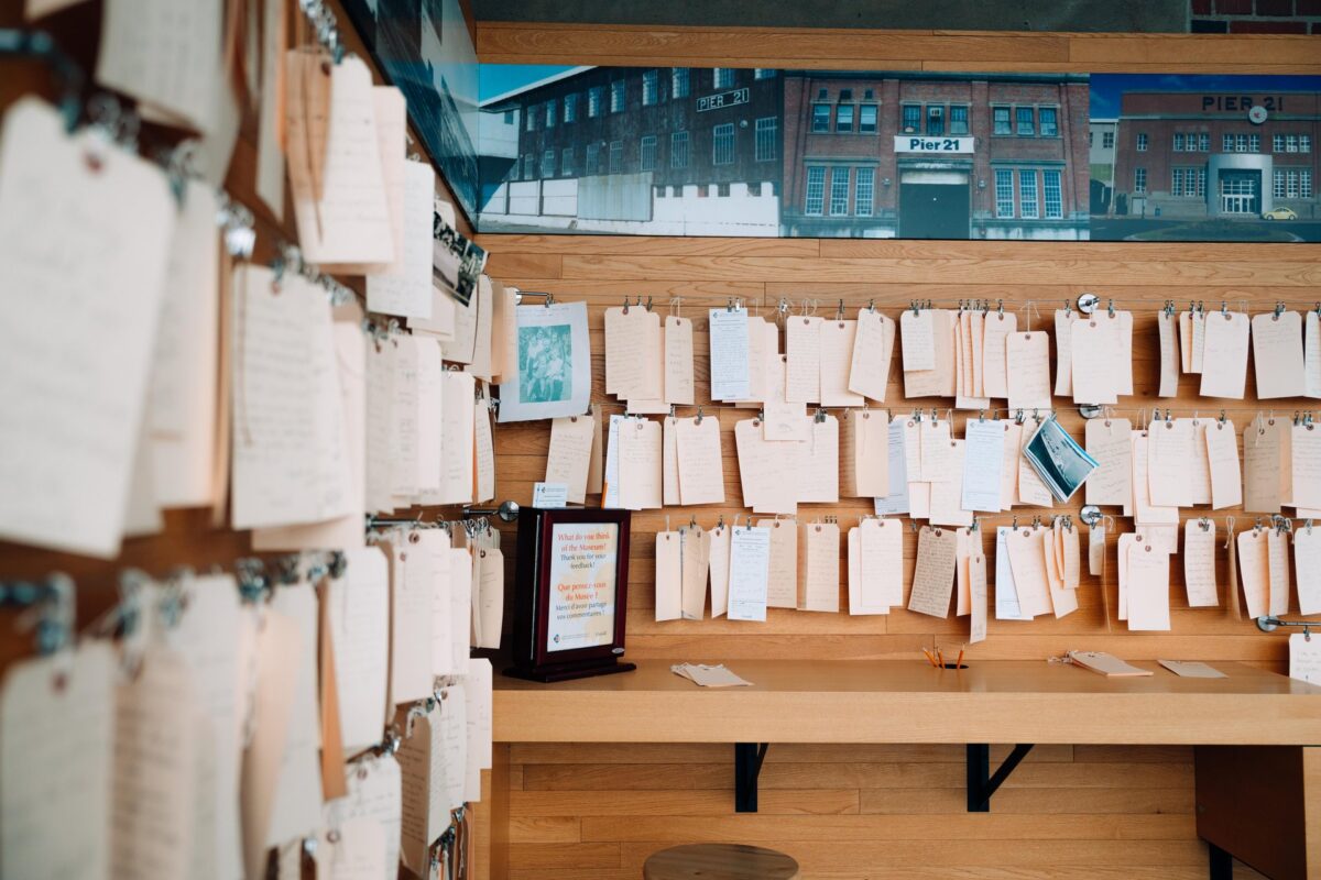 A wall of handwritten notes and papers clipped to strings at the Canadian Museum of Immigration at Pier 21, with the Pier 21 building visible in a large photo above.