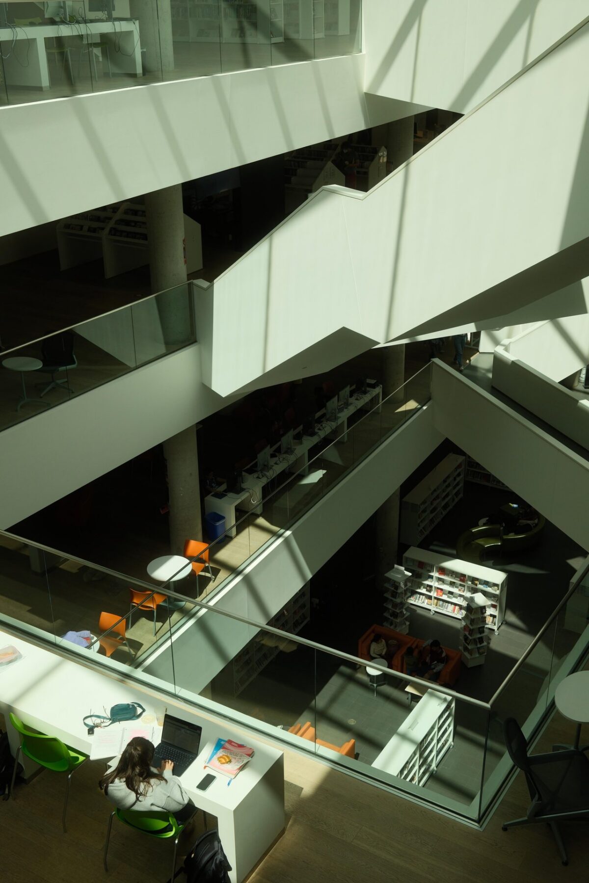 An overhead view of Halifax Central Library’s bright, multi-level interior, with glass railings, white staircases, reading areas, and sunlight casting shadows across the space.