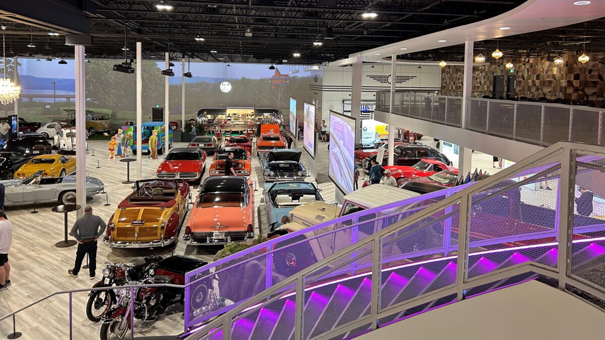 A large collection of vintage cars and motorcycles displayed inside Steele Wheels Motor Museum, viewed from an upper-level walkway with purple lighting.